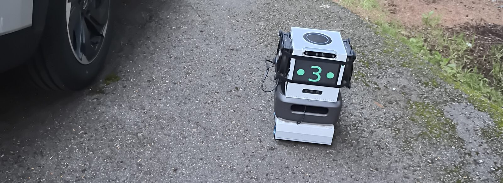 The Neural Cube robot on a gravel road at a forest campsite, next to a car wheel